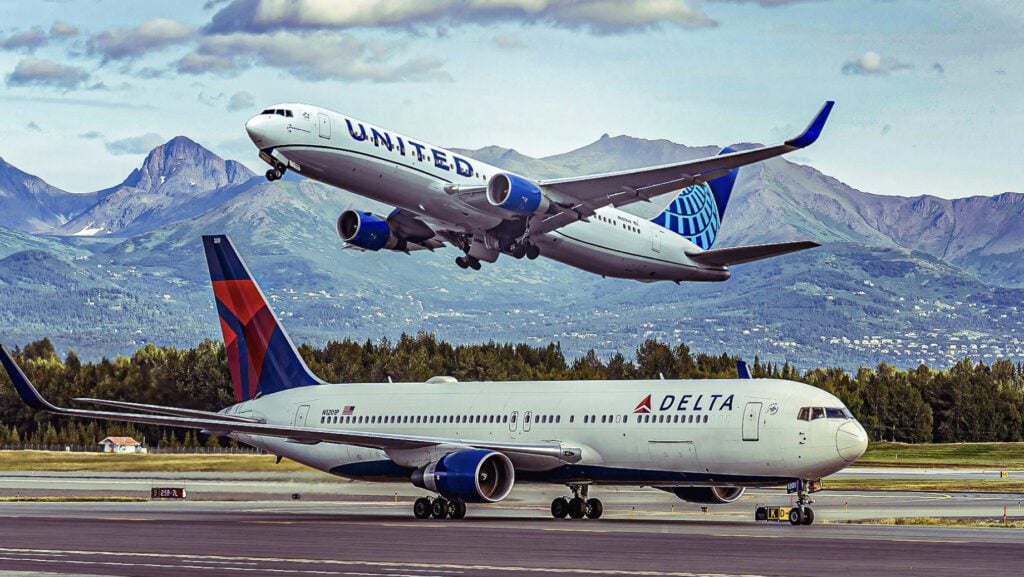 United Airlines jet lifting off the runway as a Delta airplane sits on the ground, with mountains in the background on a clear day.