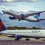 United Airlines jet lifting off the runway as a Delta airplane sits on the ground, with mountains in the background on a clear day.